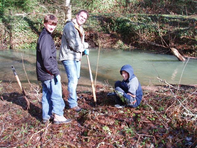Three cub scounts around the river. The older guy is propped up with a stick.